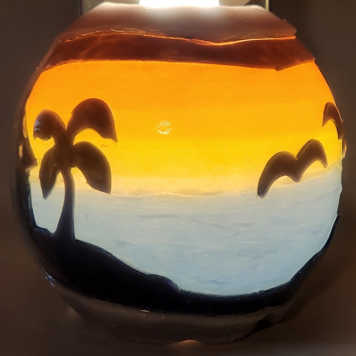 Hand carved spherical candle with Colorful sunset over blue ocean and silhouetted palm trees featured. It is lit from the top by a white light and the candle glows from within.