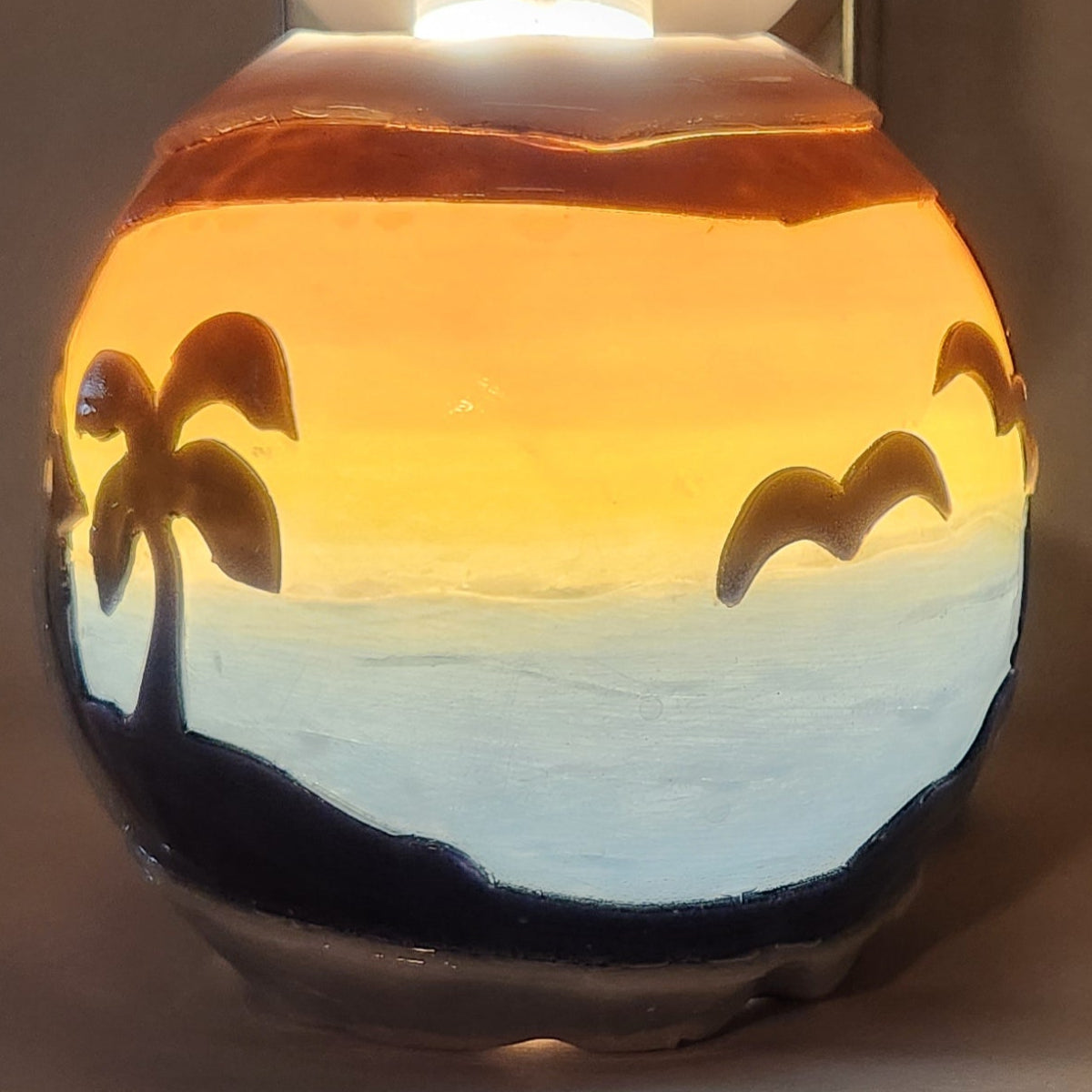 Hand carved spherical candle with colorful sunset over blue ocean and silhouetted palm trees featured. It is lit from the top by a white light and the candle glows from within.