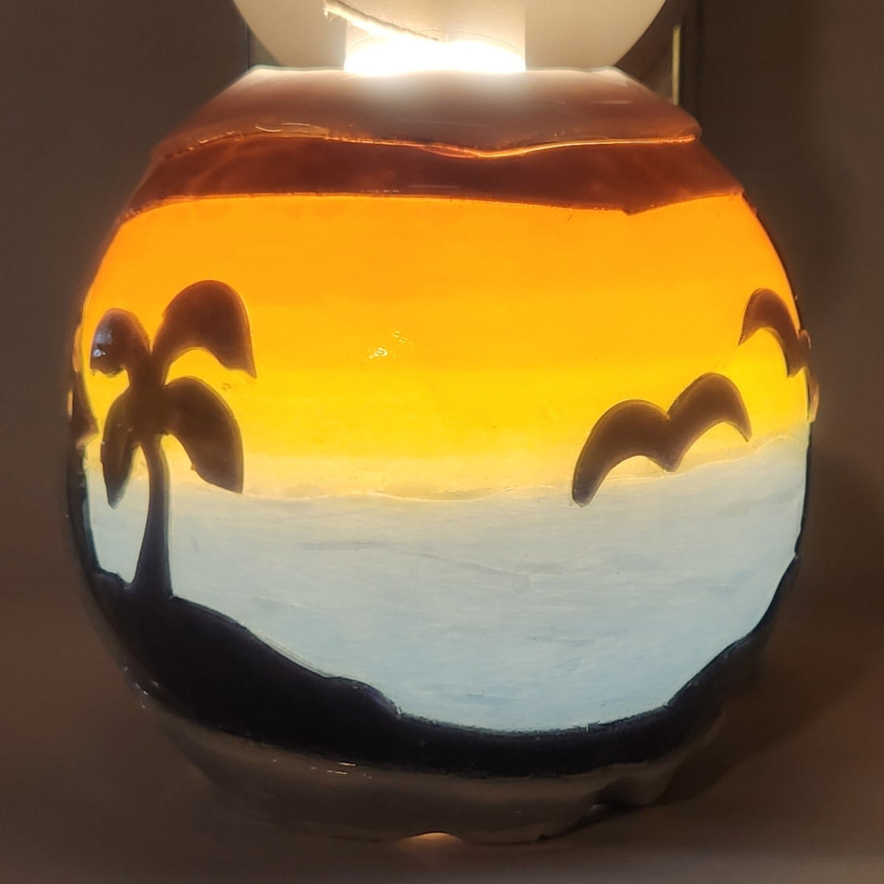 Hand carved spherical candle with colorful sunset over blue oceran and silhouetted palm trees featured. It is lit from the top by a white light and the candle glows from within.