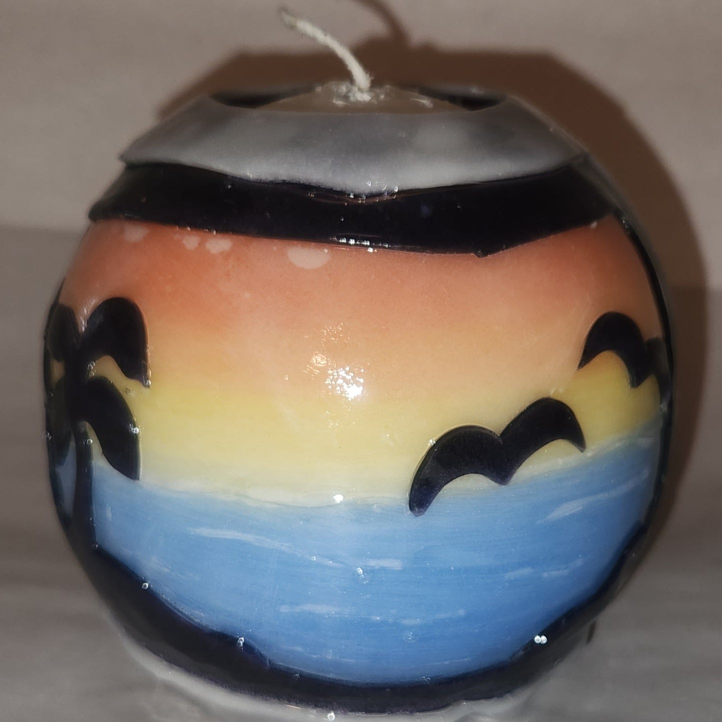 Hand carved spherical candle with colorful sunset over blue water and silhouetted palm trees featured. It is shown being lit externally.