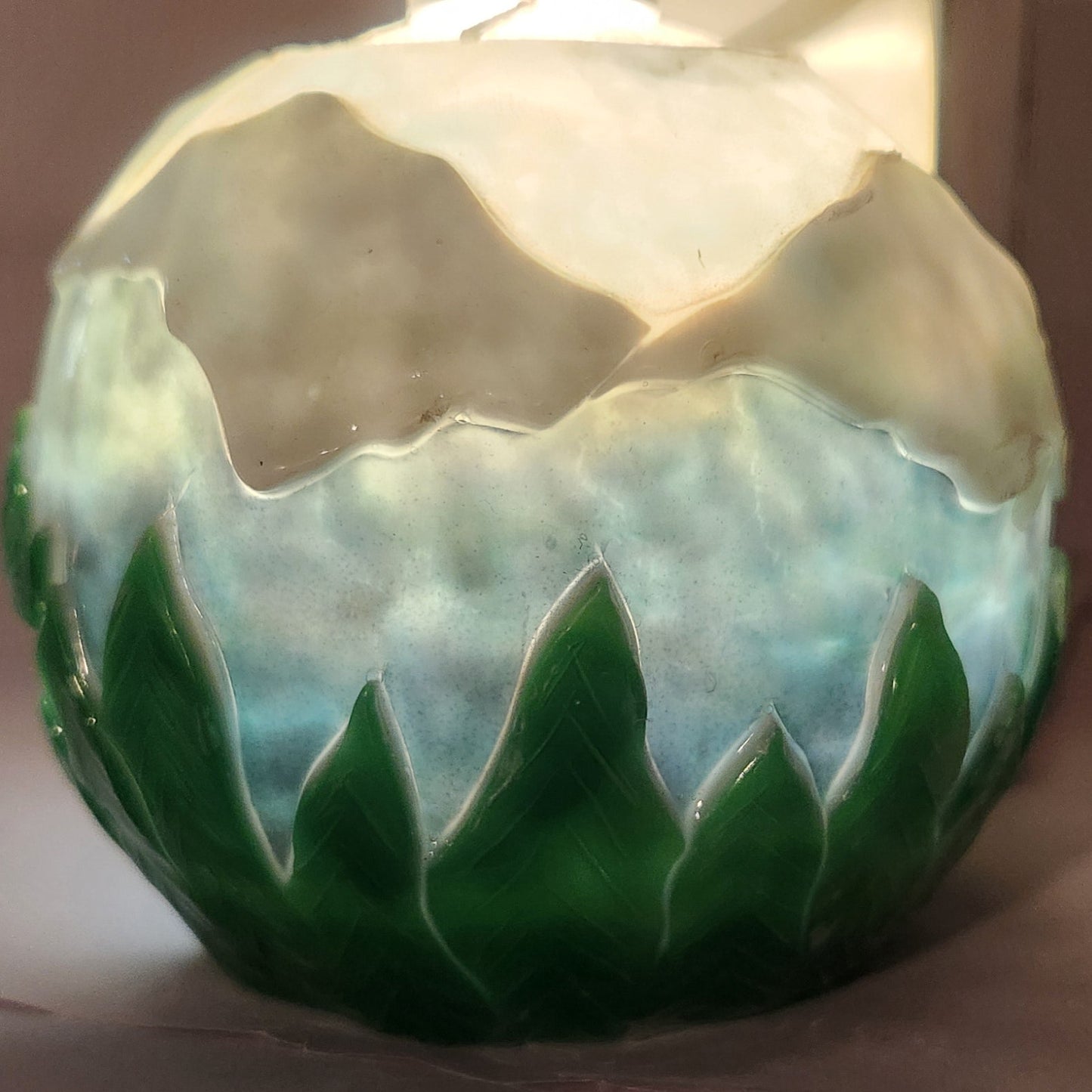 Hand carved spherical candle with trees, mountains and sky featured. It is lit from the top by a white light and the candle glows from within.