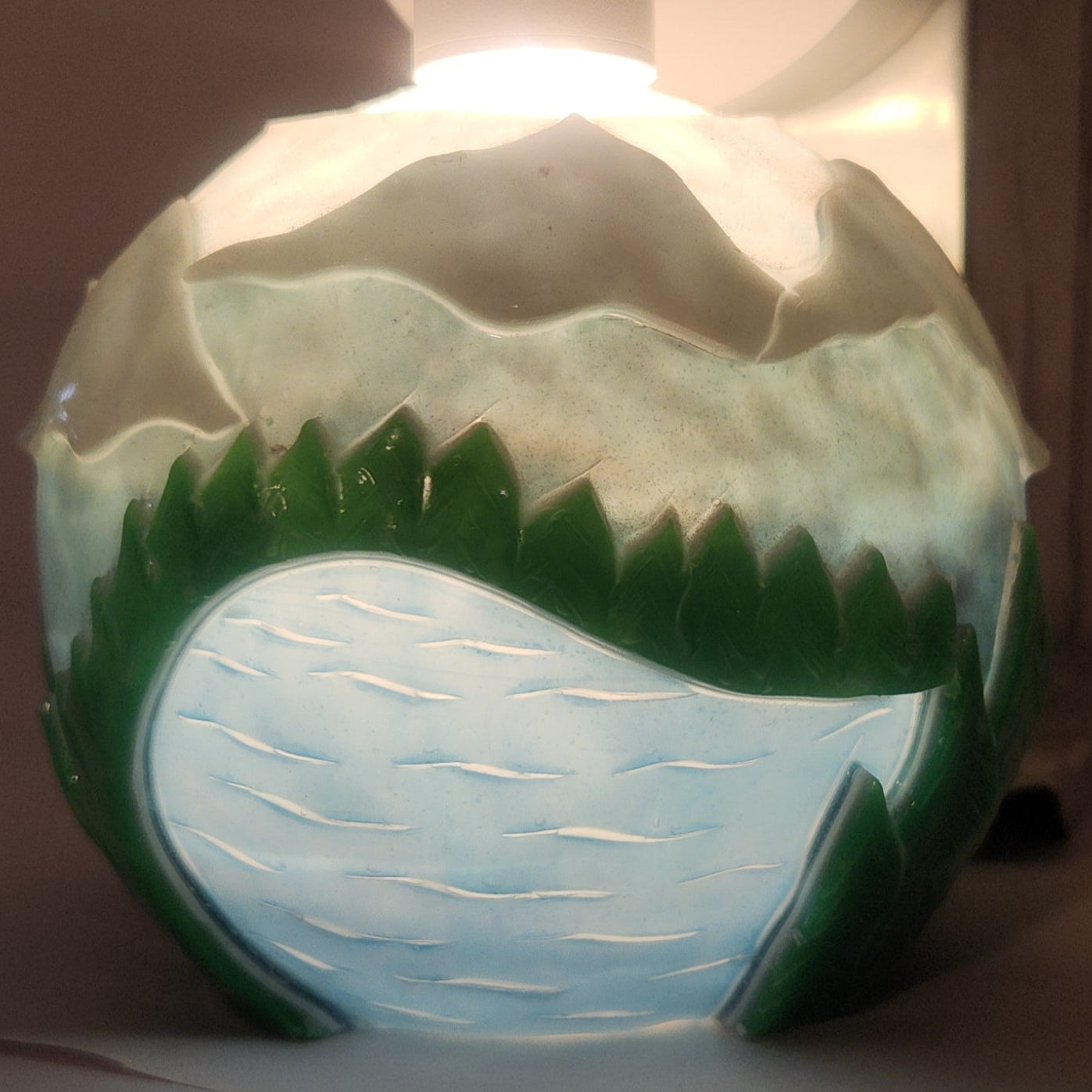 Hand carved spherical candle with rippling lake, trees, mountains and sky featured. It is lit from the top by a white light and the candle glows from within.