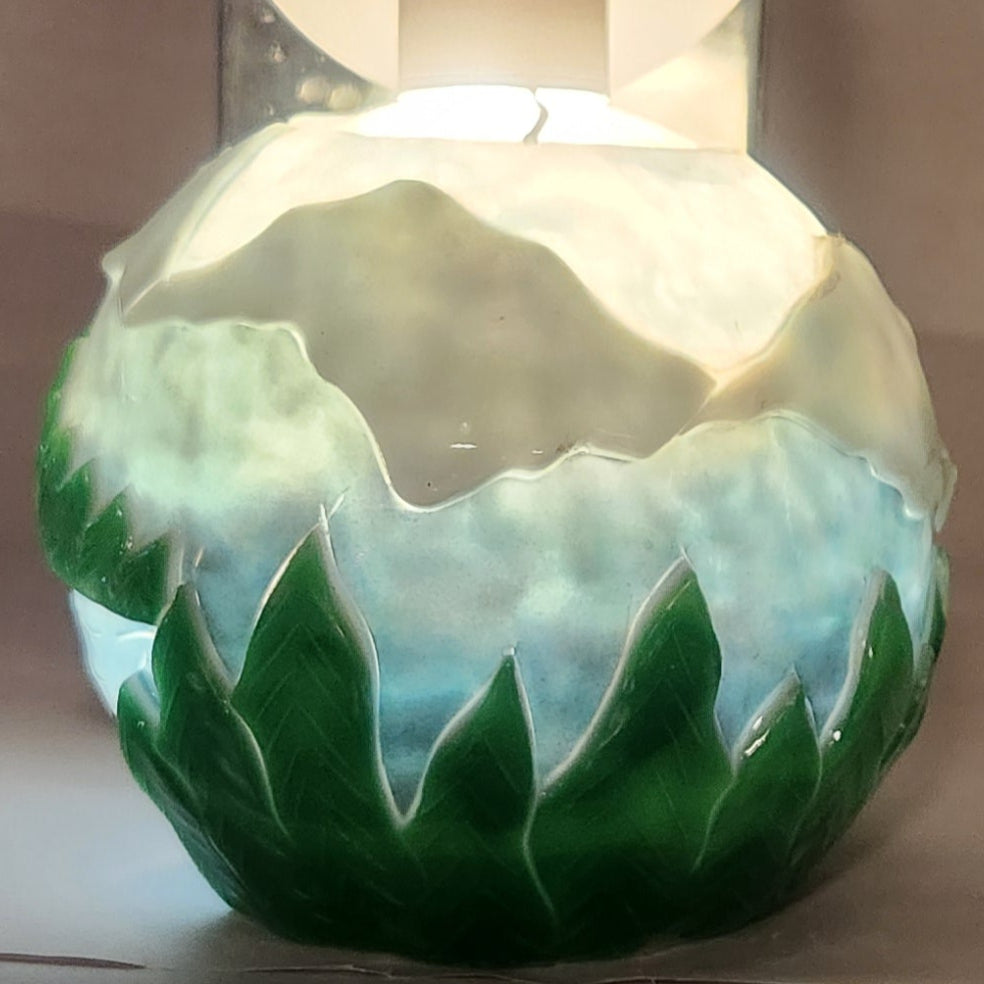 Hand carved spherical candle with trees, mountains and sky featured. It is lit from the top by a white light and the candle glows from within.