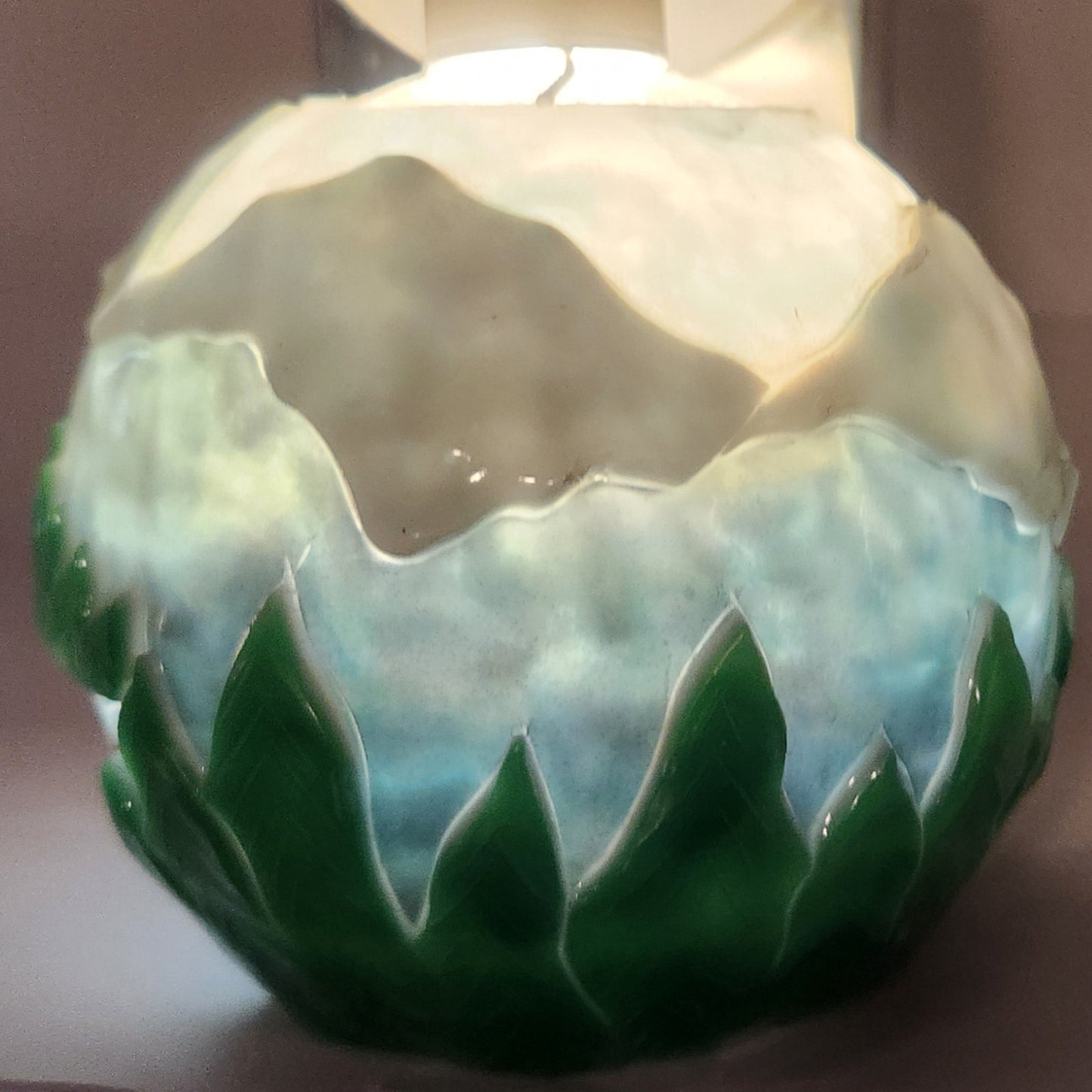 Hand carved spherical candle with trees, mountains and sky featured. It is lit from the top by a white light and the candle glows from within.