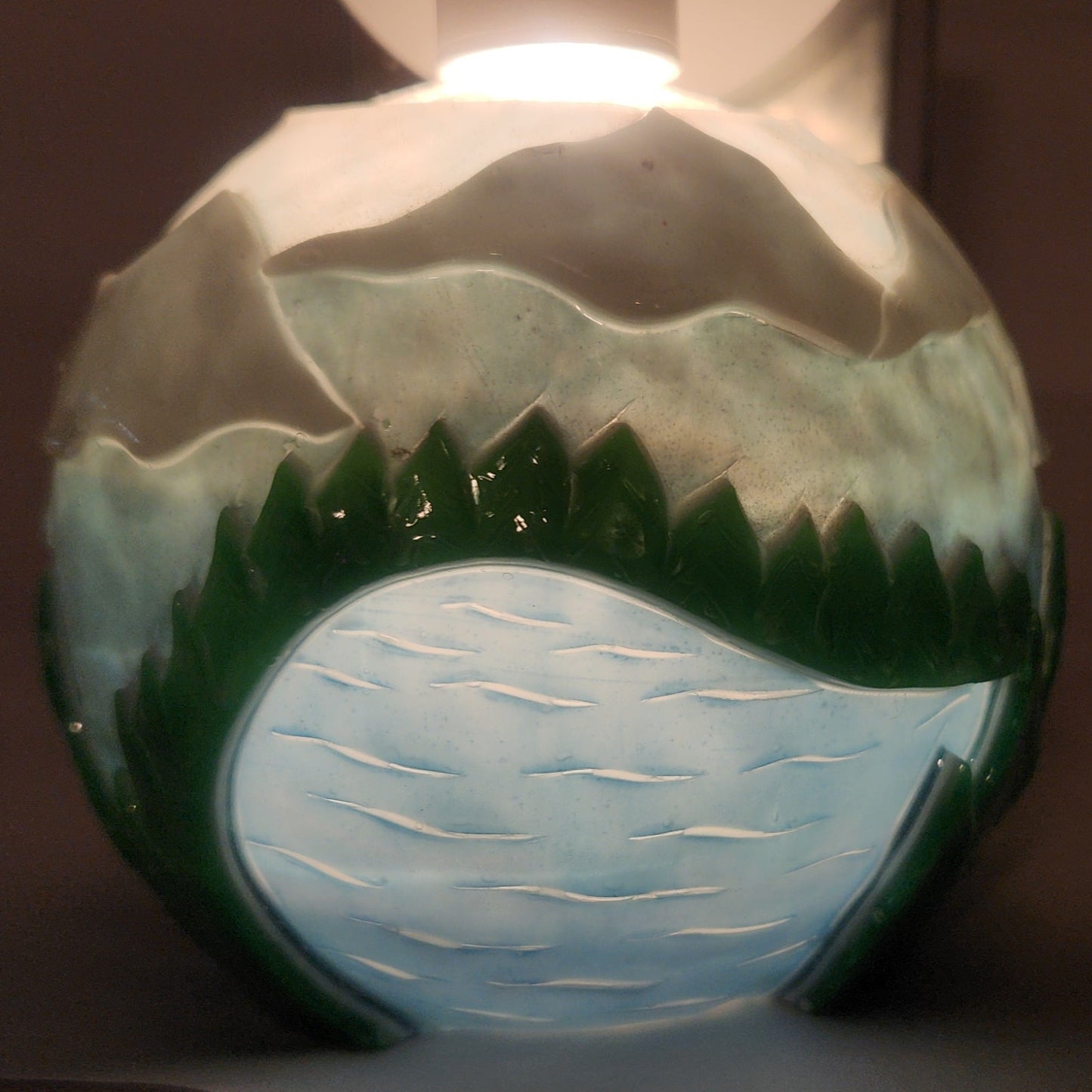 Hand carved spherical candle with rippling lake, trees, mountains and sky featured. It is lit from the top by a white light and the candle glows from within.