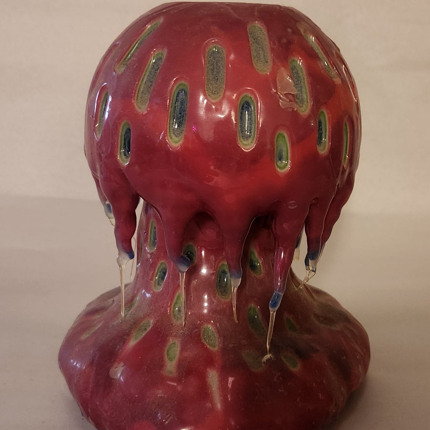 Hand carved Mushroom shaped candle with red outside and layered spots carved from the cap and stem with wax drips coming off the cap featured. It is shown lit externally.