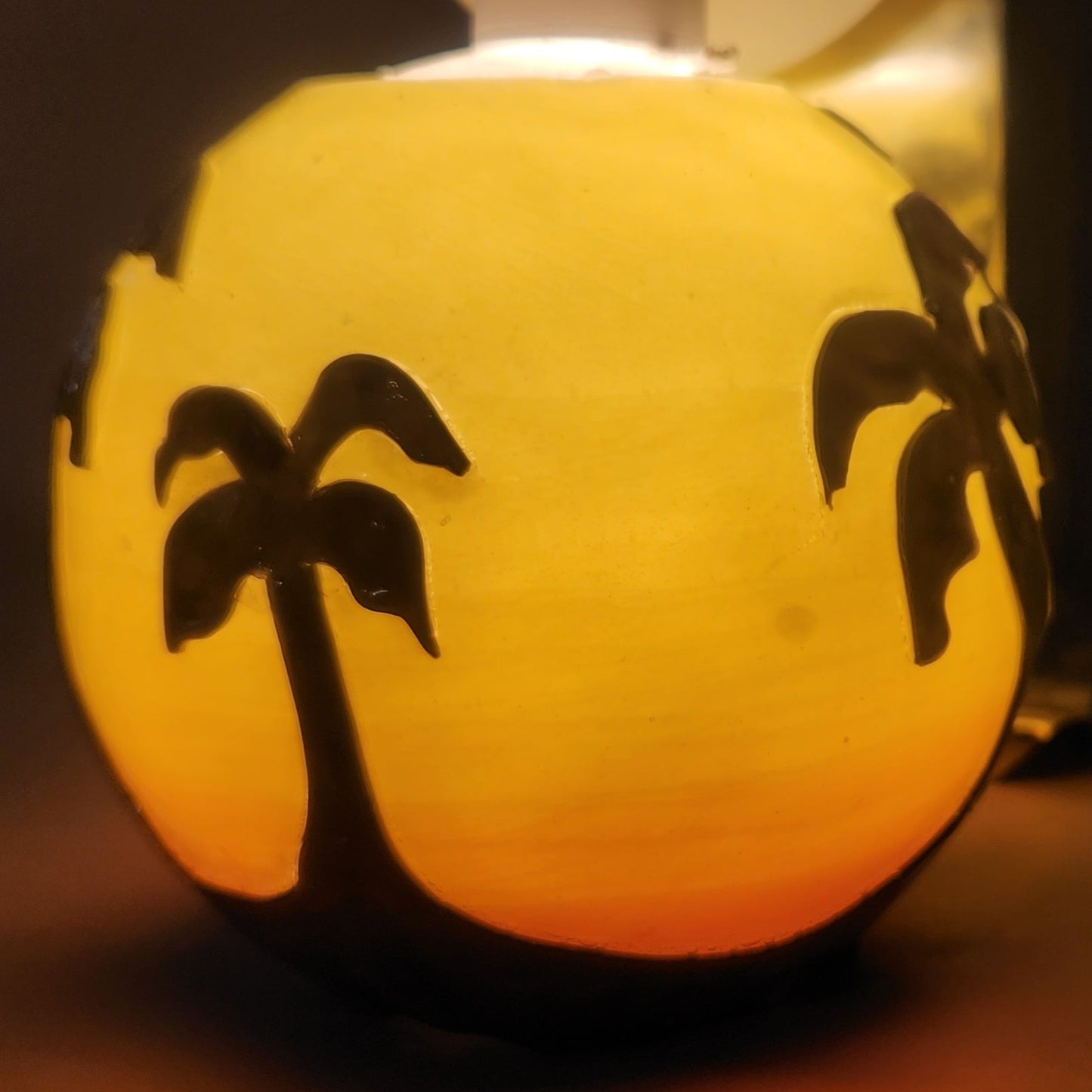 Hand carved spherical candle with Orange sunset and silhouetted palm trees featured. It is lit from the top by a white light and the candle glows from within.