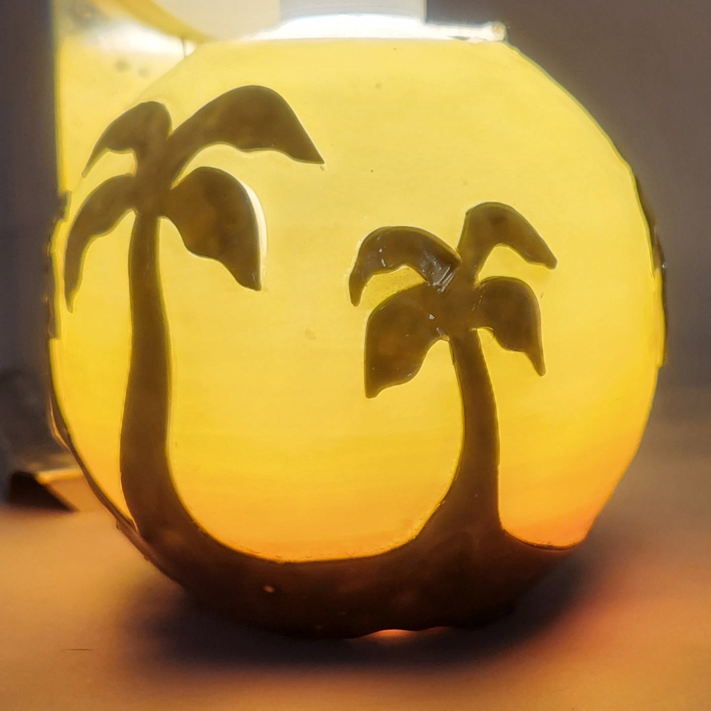 Hand carved spherical candle with Orange sunset and silhouetted palm trees featured. It is lit from the top by a white light and the candle glows from within.