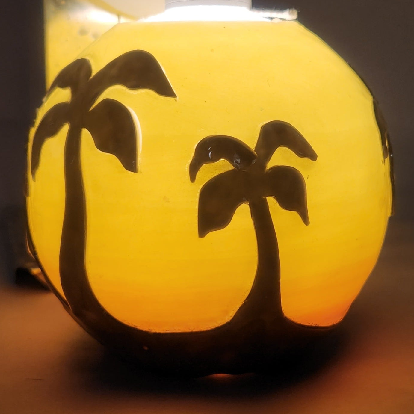 Hand carved spherical candle with Orange sunset and silhouetted palm trees featured. It is lit from the top by a white light and the candle glows from within.