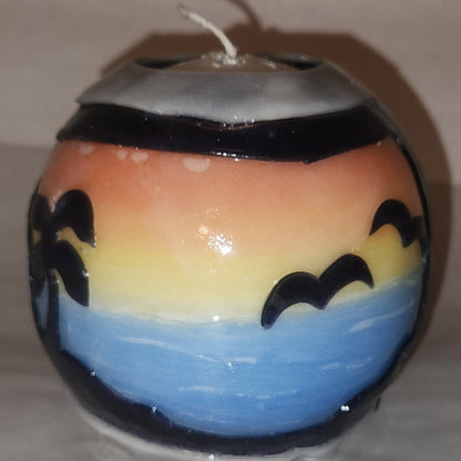 Hand carved spherical candle with colorful sunset over blue water and silhouetted palm trees featured. It is shown being lit externally.