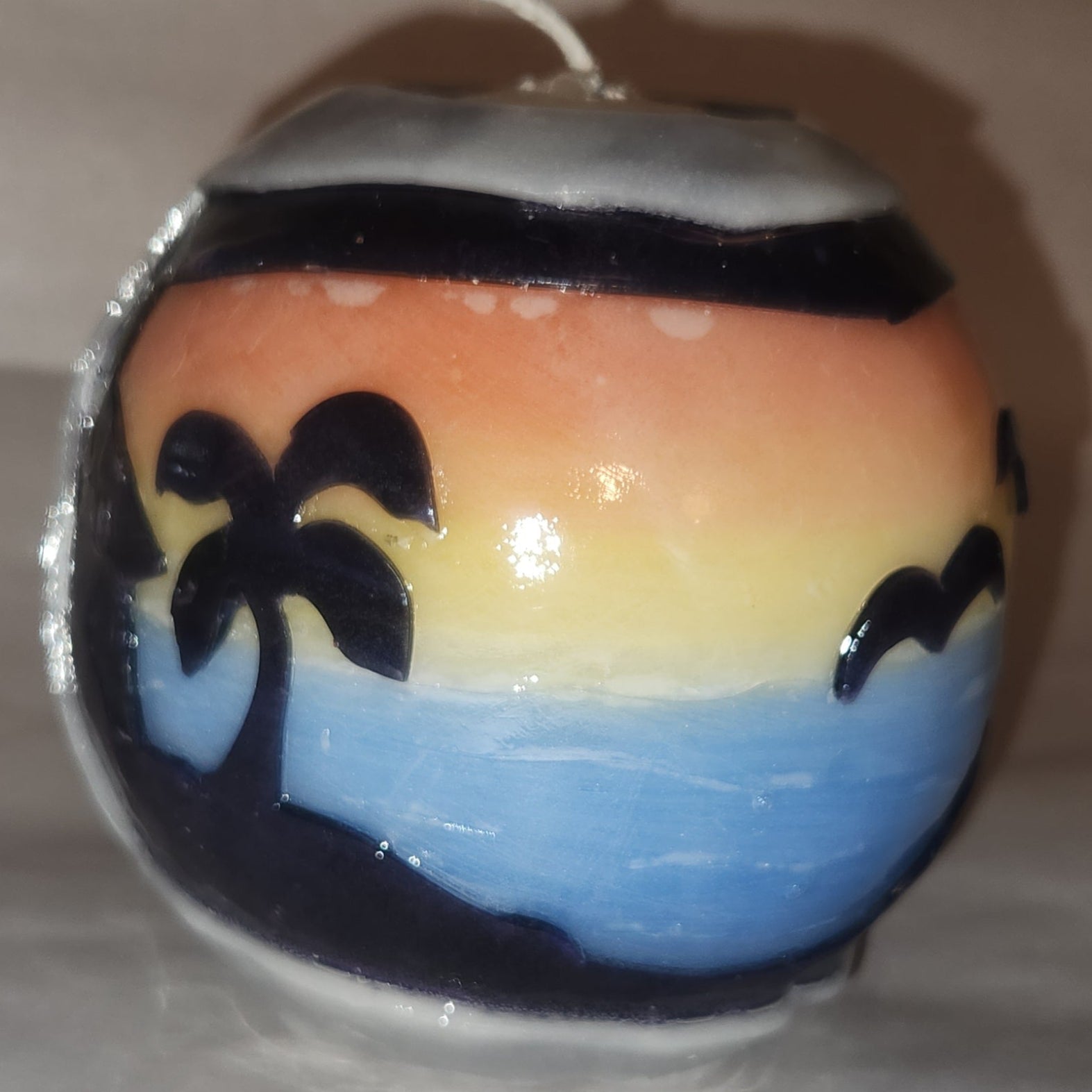 Hand carved spherical candle with colorful sunset over blue ocean and silhouetted palm trees featured. Shown lit externally.