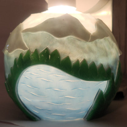 Hand carved spherical candle with rippling lake, trees, mountains and sky featured. It is lit from the top by a white light and the candle glows from within.