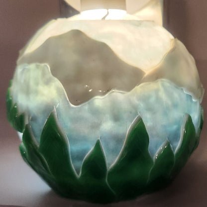 Hand carved spherical candle with trees, mountains and sky featured. It is lit from the top by a white light and the candle glows from within.