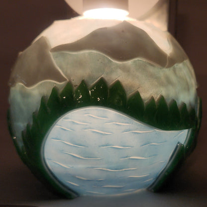 Hand carved spherical candle with rippling lake, trees, mountains and sky featured. It is lit from the top by a white light and the candle glows from within.