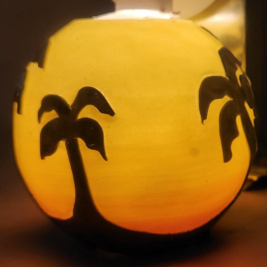 Hand carved spherical candle with Orange sunset and silhouetted palm trees featured. It is lit from the top by a white light and the candle glows from within.